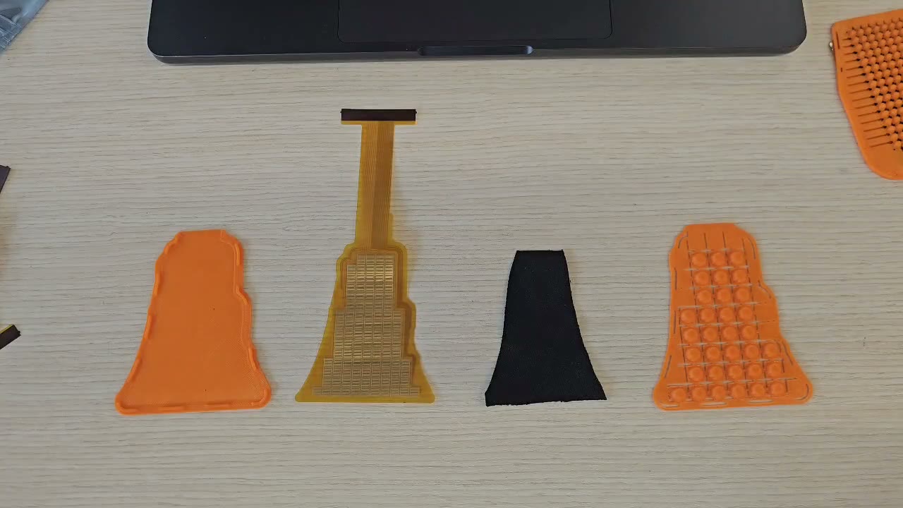 The parts of one sensor laid out on a desk: the printed TPU tray, the flex PCB with its tail, the black piezoresistive textile cut to shape, and the printed bump sheet.